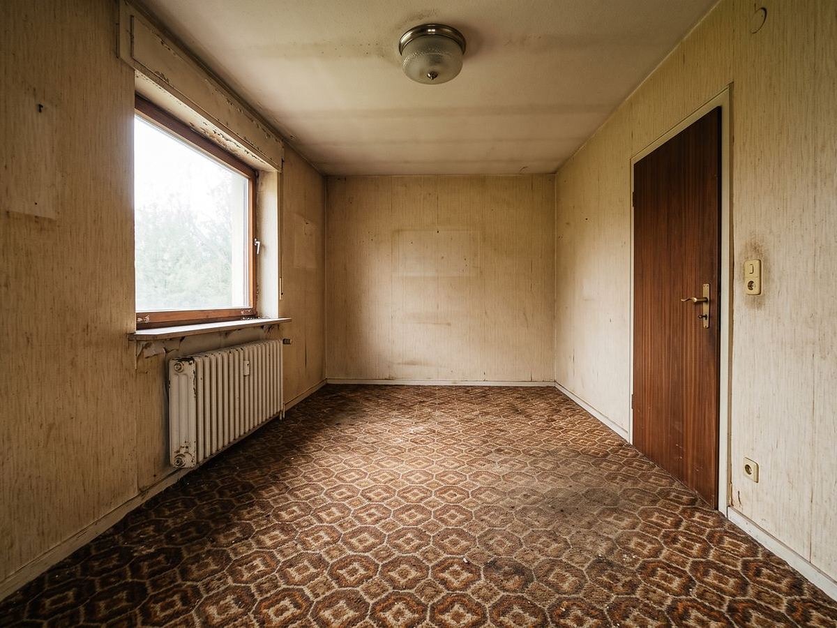 Vorher: Wohnraum mit gemustertem Teppichboden, Tapete und altem Heizkörper